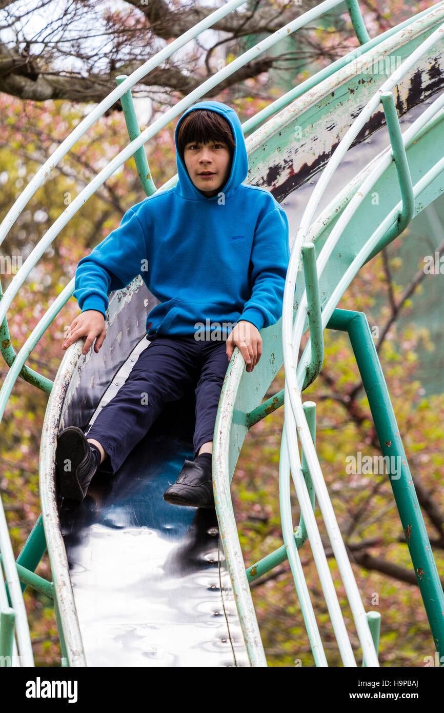 Japan, Akashi. Kazki, early teenage caucasian boy, 13-14 years old, in blue hoodie sliding on curved slide towards viewer. Eye-contact. Stock Photo