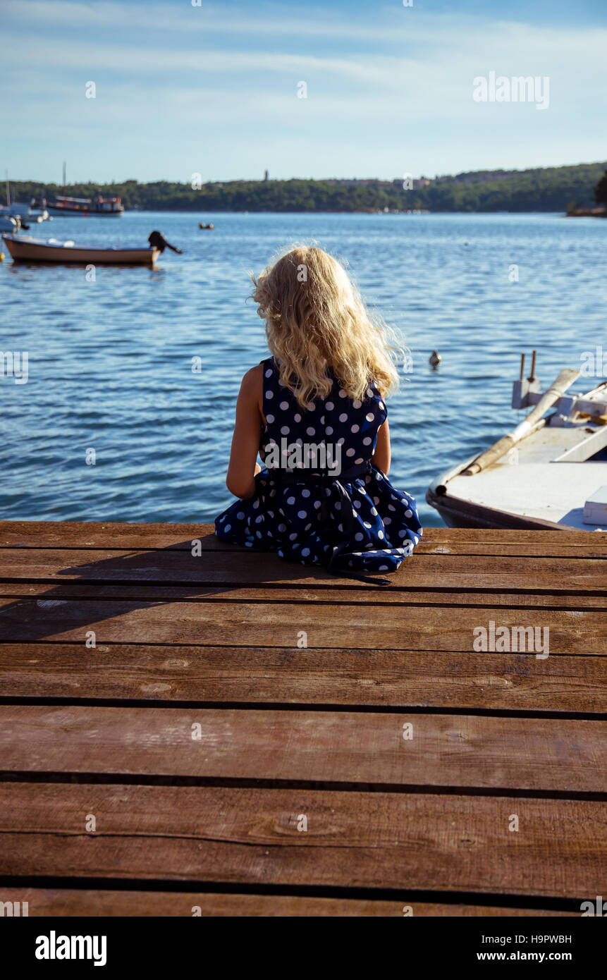little girl with long blond hair sitting on the edge of wooden jetty Stock Photo
