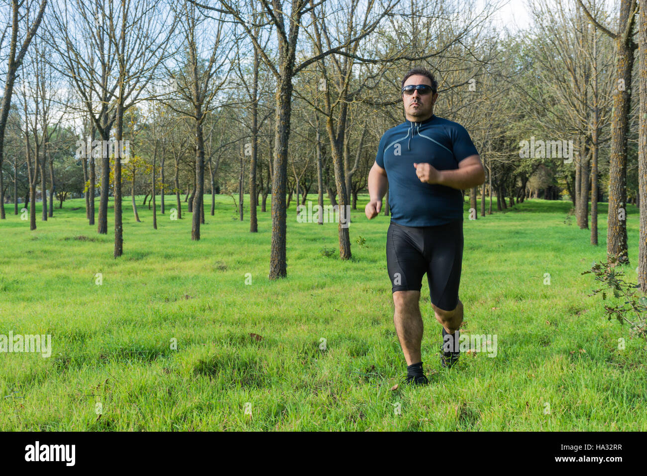 Big belly man jogging , exercising, doing cardio in the park , slightly overweight, loosing weight. On a lawn of green grass between trees without lea Stock Photo