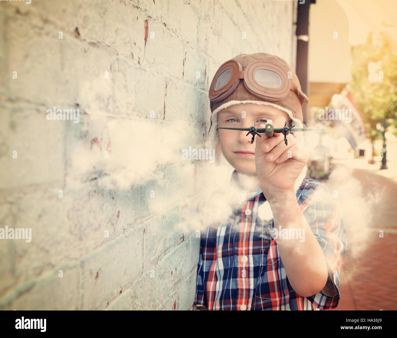 A young boy is pretending to be a pilot and playing with an airplane toy against a brick wall for a dream or career concept. Stock Photo