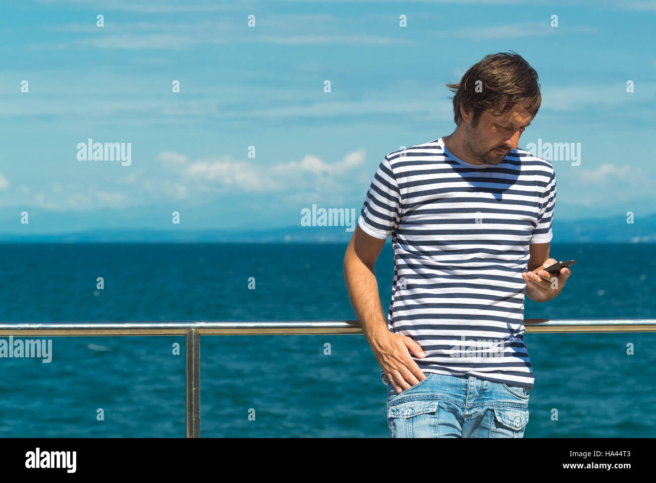 Man in striped sailor shirt sending SMS by the sea. Using mobile phone for communication on summertime beach holiday vacation. Stock Photo