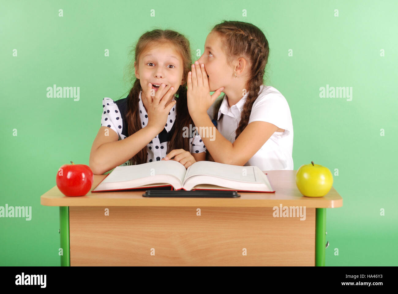 Two school girl sharing secrets  sitting at a desk from book isolated on green Stock Photo