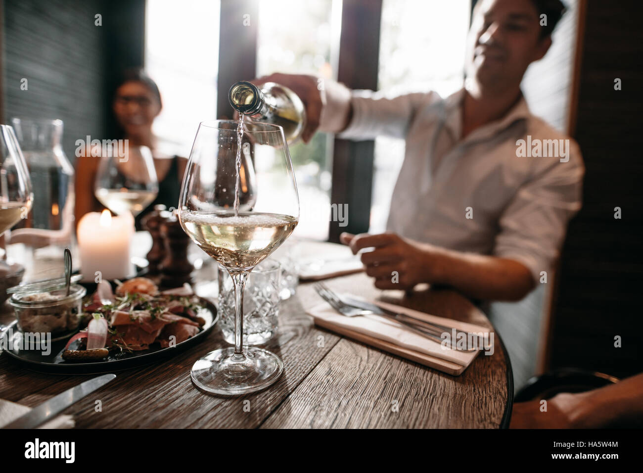 Young man pouring wine from the bottle into a glass with friends sitting around the table. Young people having wine at restaurant. Stock Photo