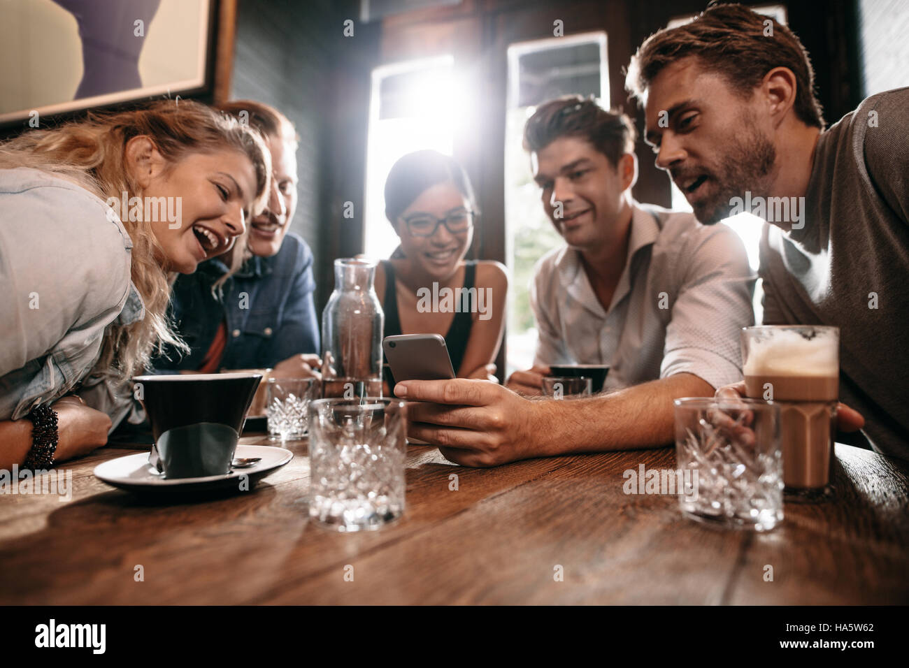 Young friends looking at smart phone while sitting in cafe. Mixed race people sitting at a table in restaurant using mobile phone. Stock Photo