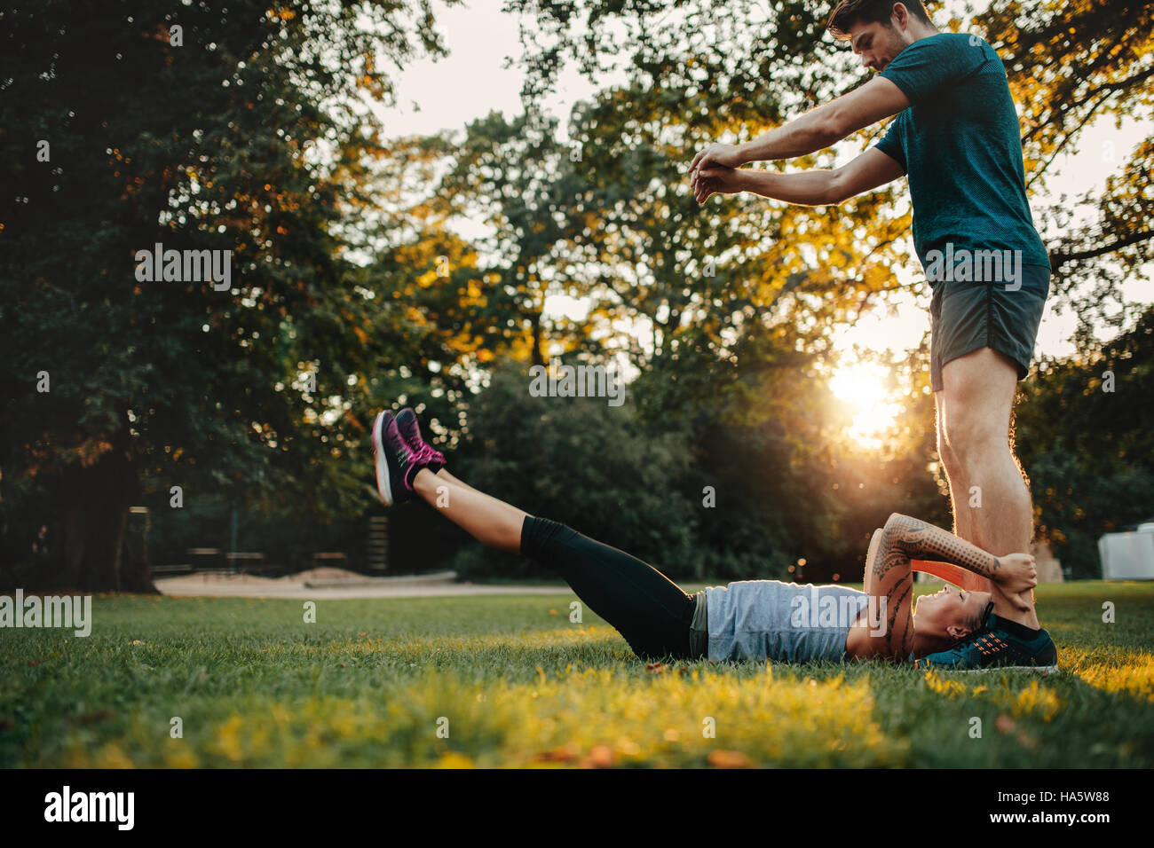 Woman lying on grass and stretching with support from her personal trainer. Couple exercising together in park. Stock Photo