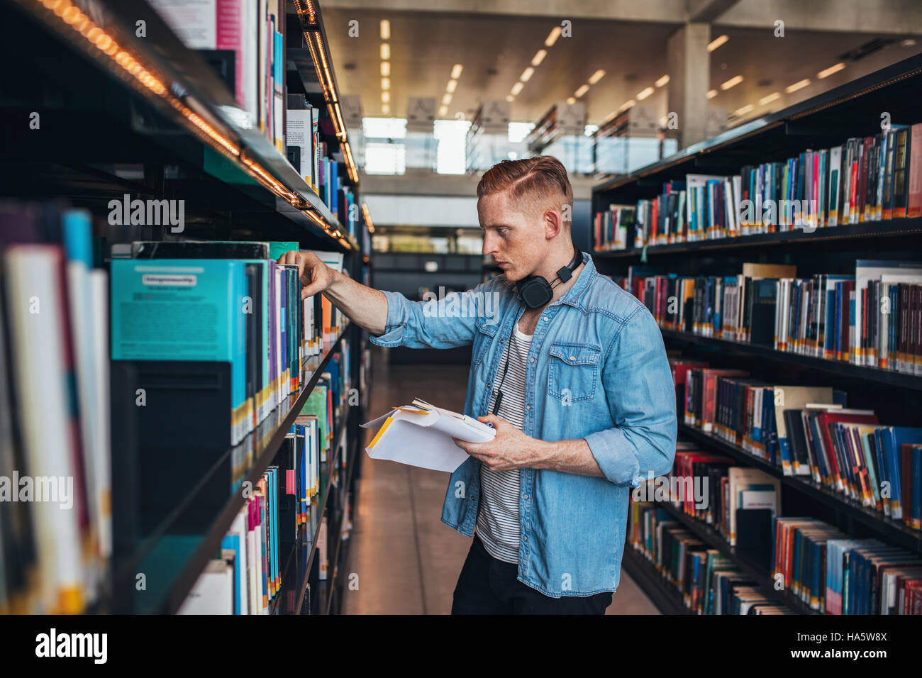 Young college student taking book from shelf in library. Handsome young caucasian man in library. Stock Photo