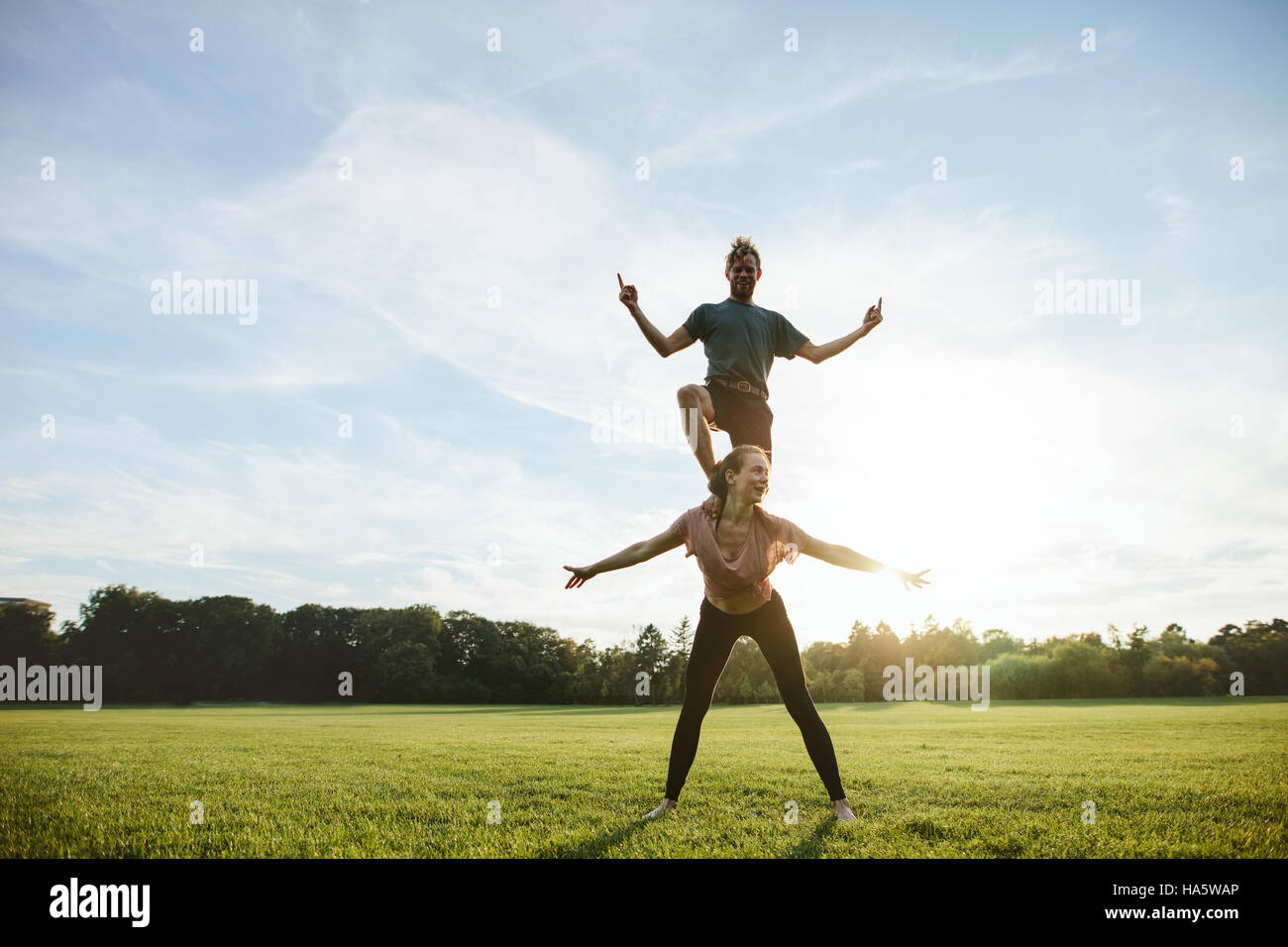 Strong young couple doing acrobatic yoga exercise in park. Man standing on back of woman and balancing. Stock Photo