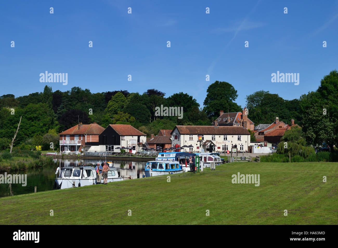 Boats moored at the staithe on the River Bure at Coltishall Common with the Rising Sun pub beyond on the Norfolk Broads. Stock Photo