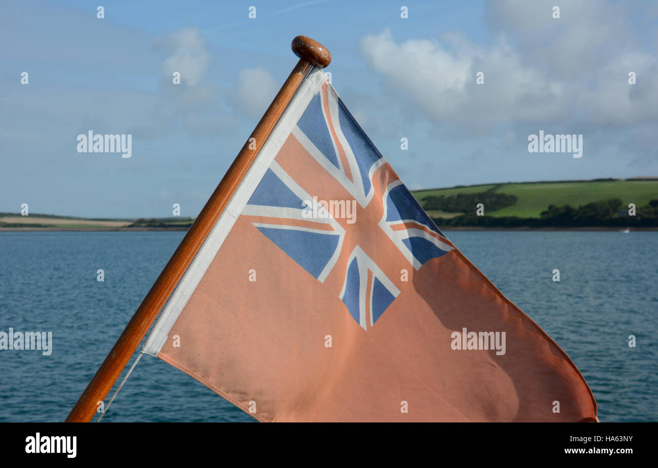 Red Ensign on yachts flag pole with blue sky & white clouds, green hills and ripple azure sea Stock Photo