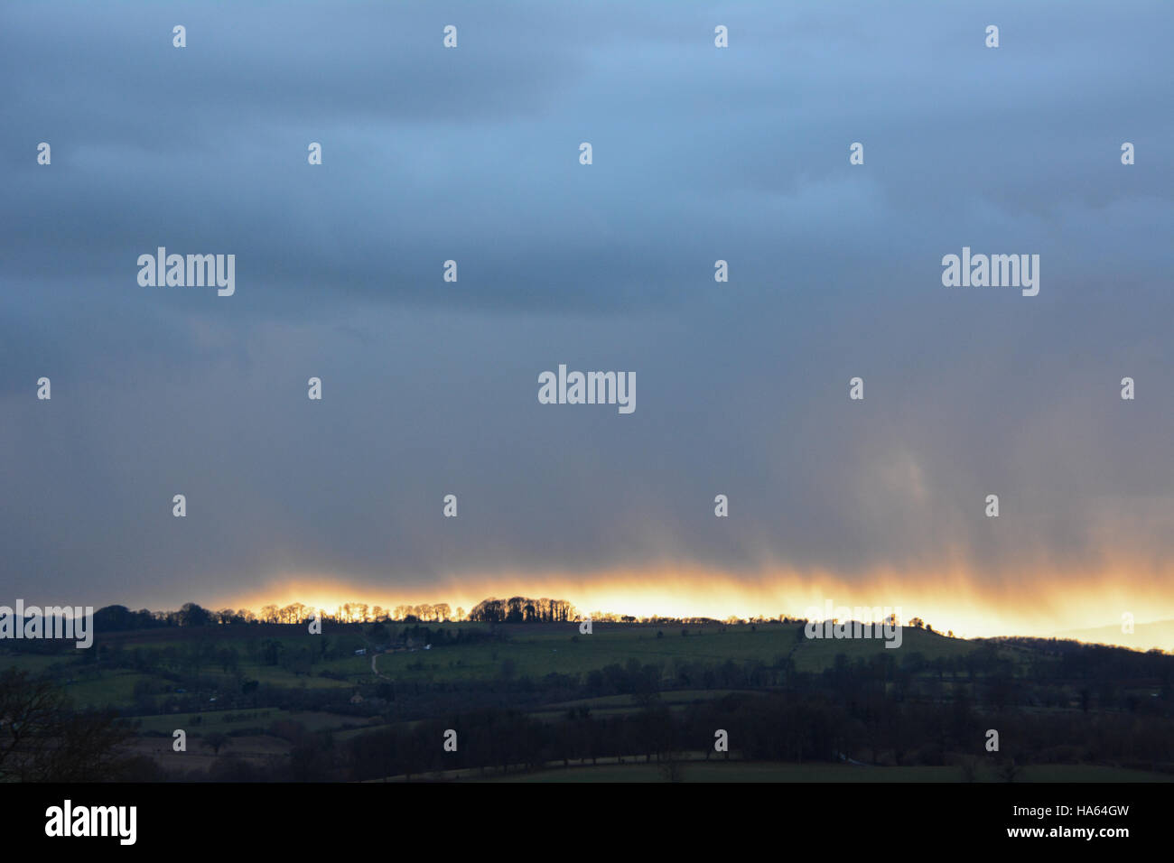 Heavy dark sky begins to drop rain over green Cotswold hills with the glow of sunset disappearing below Stock Photo