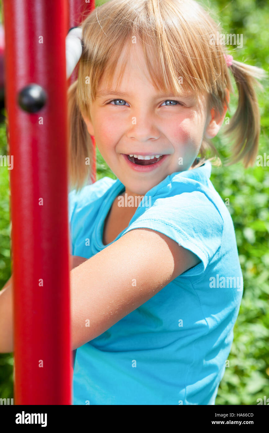 Portrait of cute blond girl with blue eyes wearing blue tshirt sitting on monkey bars on a summer day. Girl looking at camera smiling Stock Photo