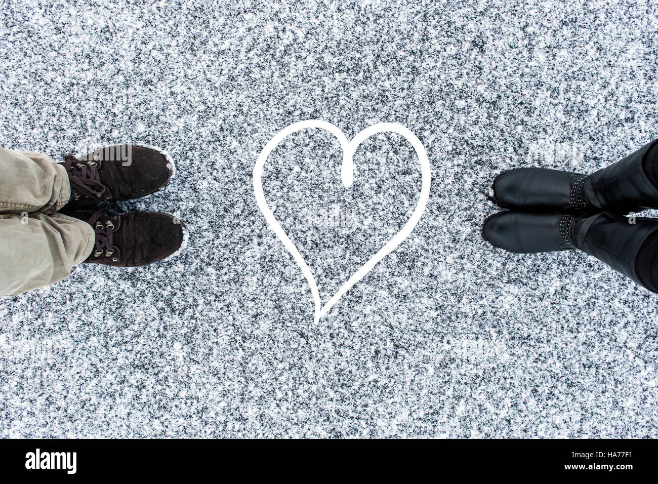 Male and Female boots standing at heart symbol on asphalt covered gritty snow surface. Rough snowy. Cold Winter. Top view. Relations concept. Stock Photo