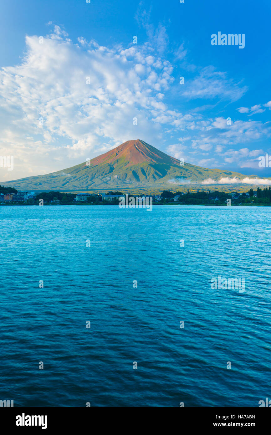 Centered Mount Fuji bathes in morning side light while Lake Kawaguchiko sits in shadow below cloudy blue sky on a summer day Stock Photo