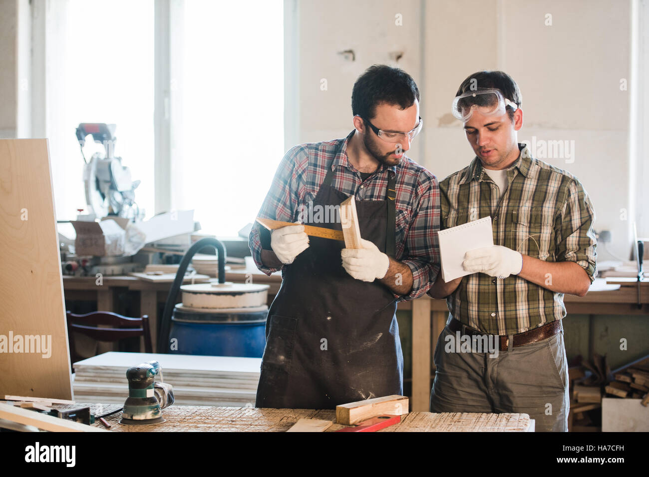 Happy male carpenter showing something to coworker at his notebook papers in workshop Stock Photo
