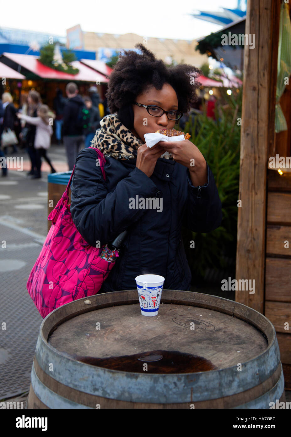 Young Lady Eating Fast Food at Hyde Park and Winter Wonderland in London she is  near a Wooden Log Cabin and is dressed in Coat Stock Photo