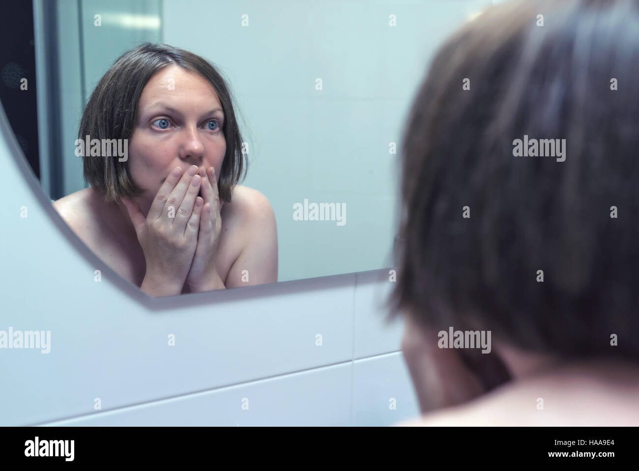 Fear of getting old, adult female in front of bathroom mirror looking at her face reflection and wrinkles on the forehead Stock Photo