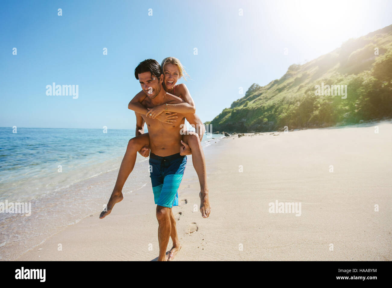 Portrait of man carrying girlfriend on his back along the sea shore. Man giving piggyback ride to girlfriend on the beach. Stock Photo