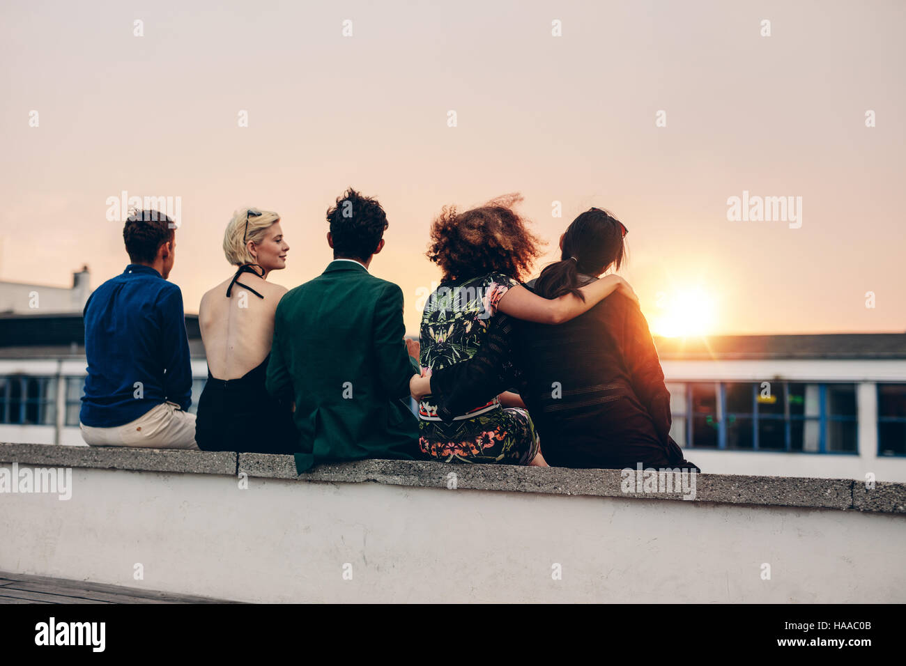 Rear view of young friends relaxing together on rooftop at sunset. Young men and women sitting on terrace in evening. Stock Photo