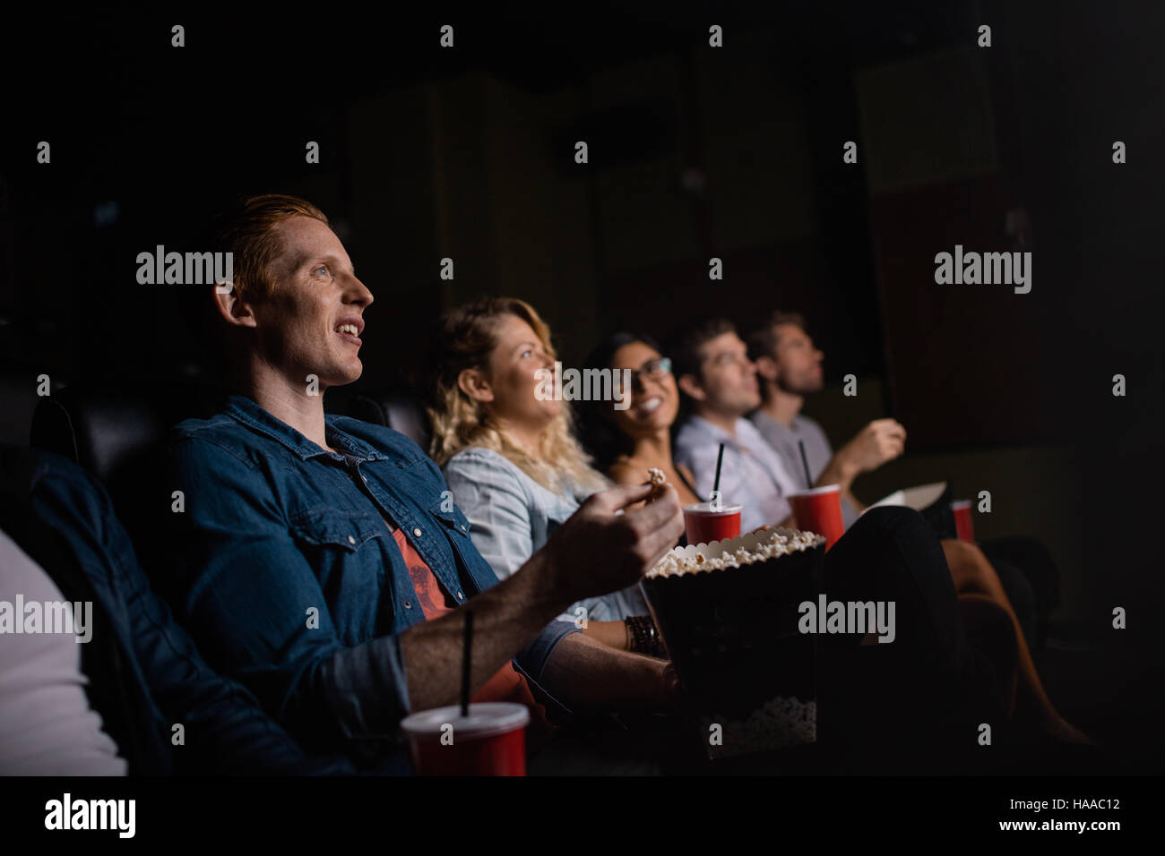 Group of friends sitting in multiplex movie theater with popcorn and drinks. Young people watching movie in cinema. Stock Photo