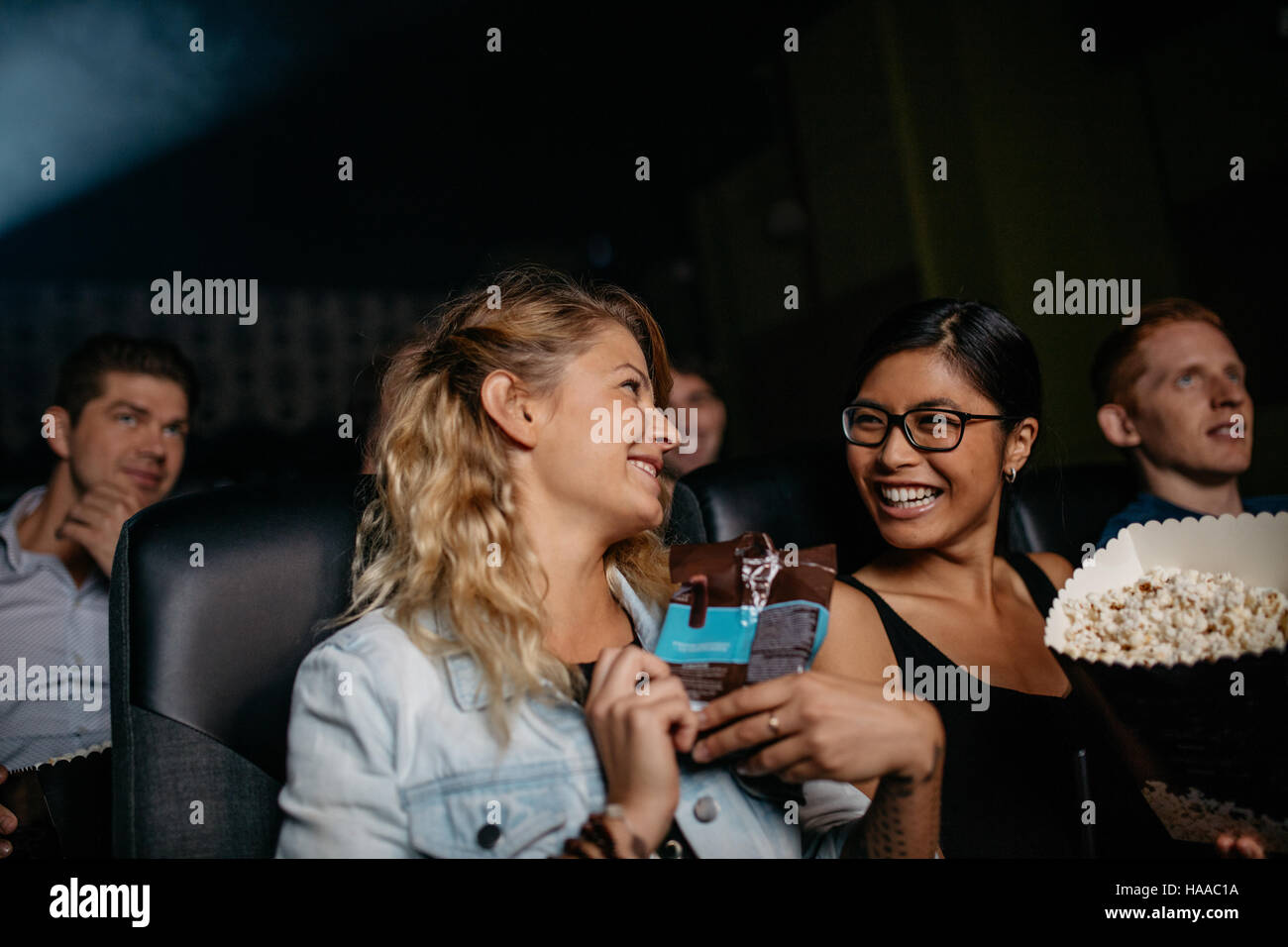 Two young woman chatting and smiling while sitting in movie theater. Group of people watching movie together. Stock Photo