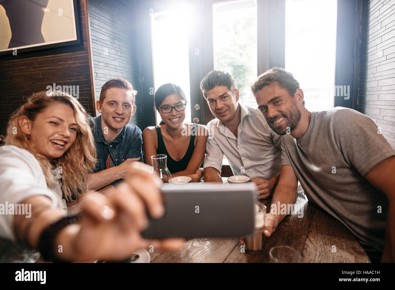 Small group of friends taking selfie on a mobile phone. Young men and women sitting together at cafe and taking a self portrait on smart phone. Stock Photo