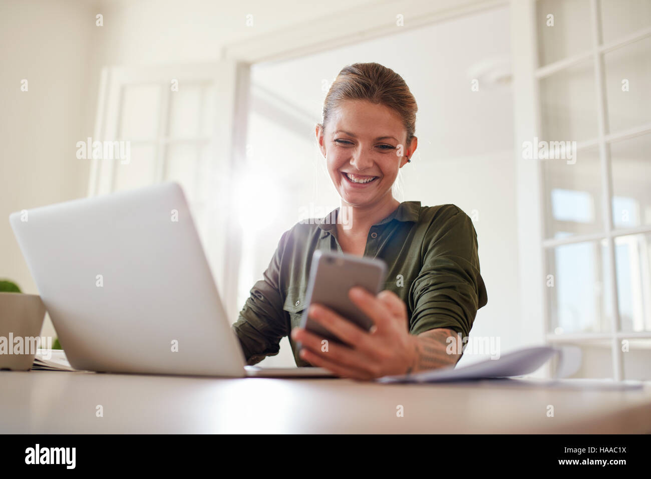 Shot of happy young woman using mobile phone while working on laptop. Female reading text message on her smart phone and smiling. Stock Photo