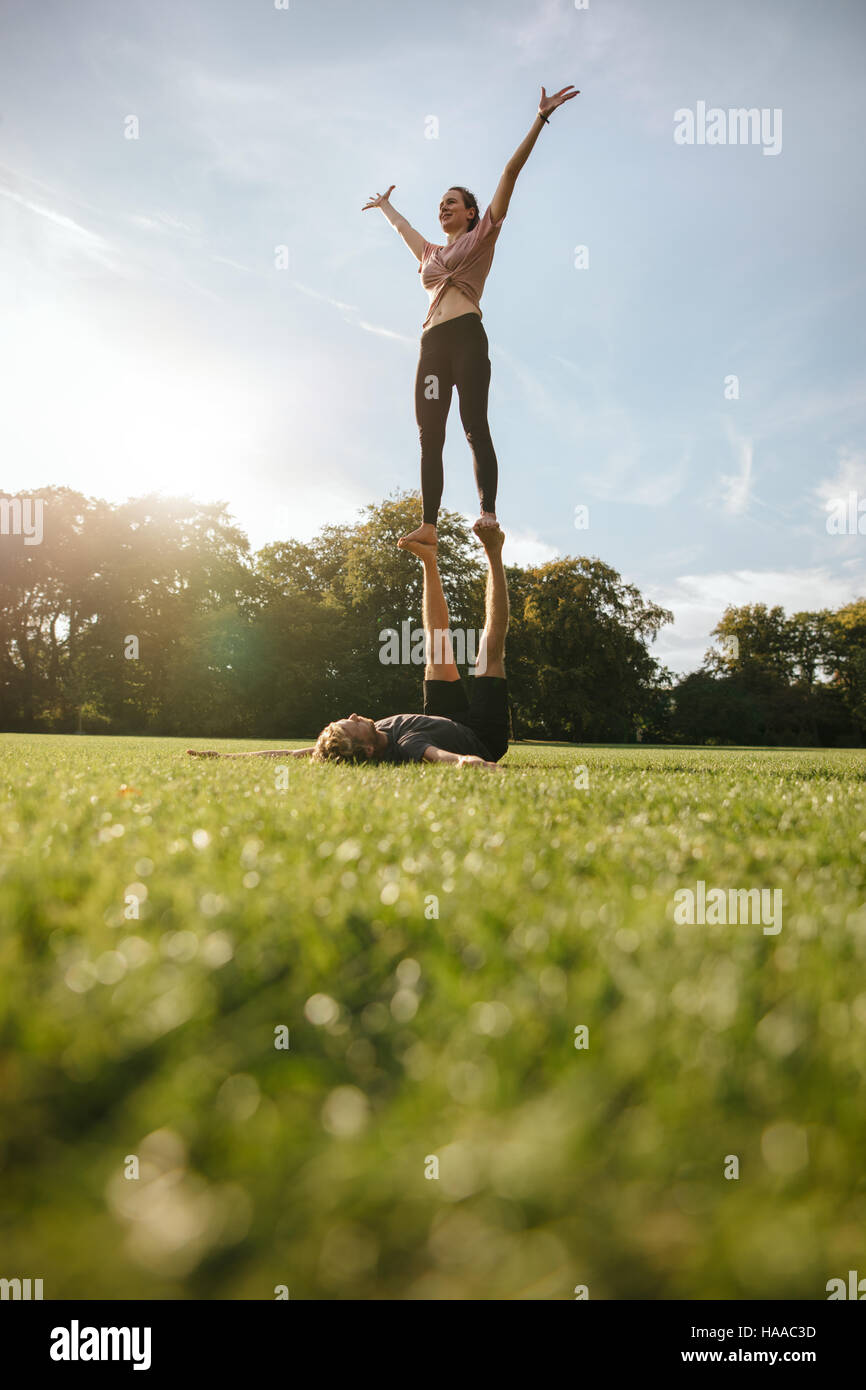 Vertical shot of fit young couple doing acroyoga exercise in park. Man lying on grass and balancing woman. Stock Photo