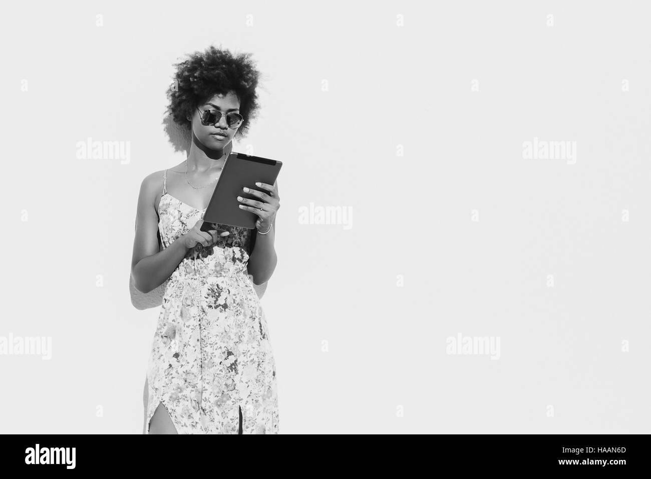 Black beautiful attractive young brazilian girl with curly hair in sunglasses and dress with digital tablet, in front of white textured wall Stock Photo