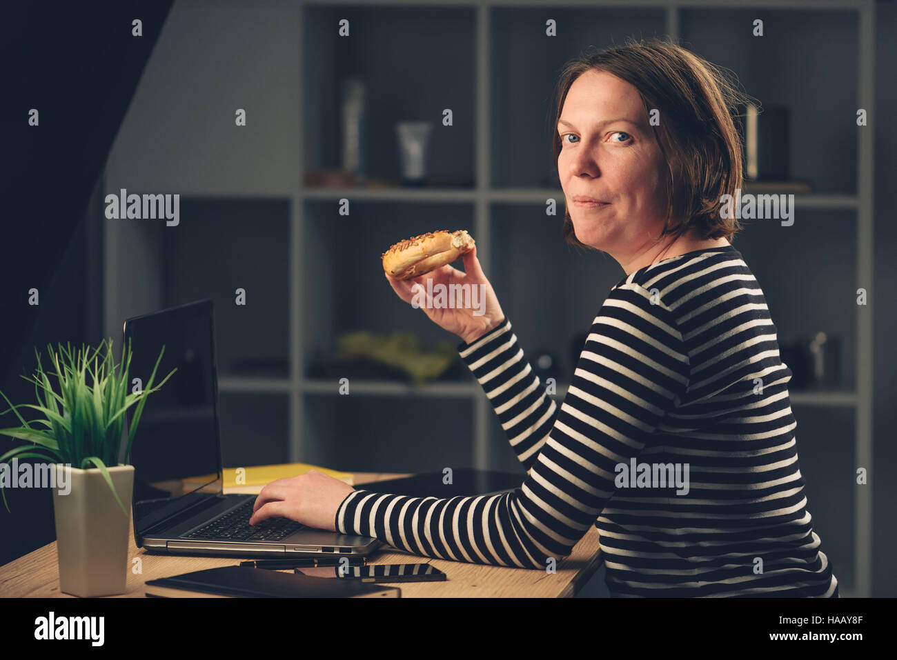Woman eating sesame bagel in office while working overtime on laptop computer Stock Photo