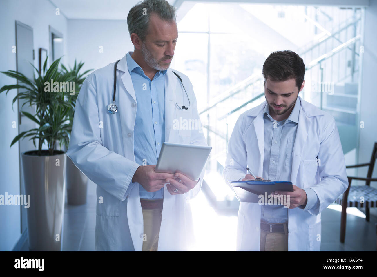 Doctors using digital tablet while writing on clipboard in corridor ...