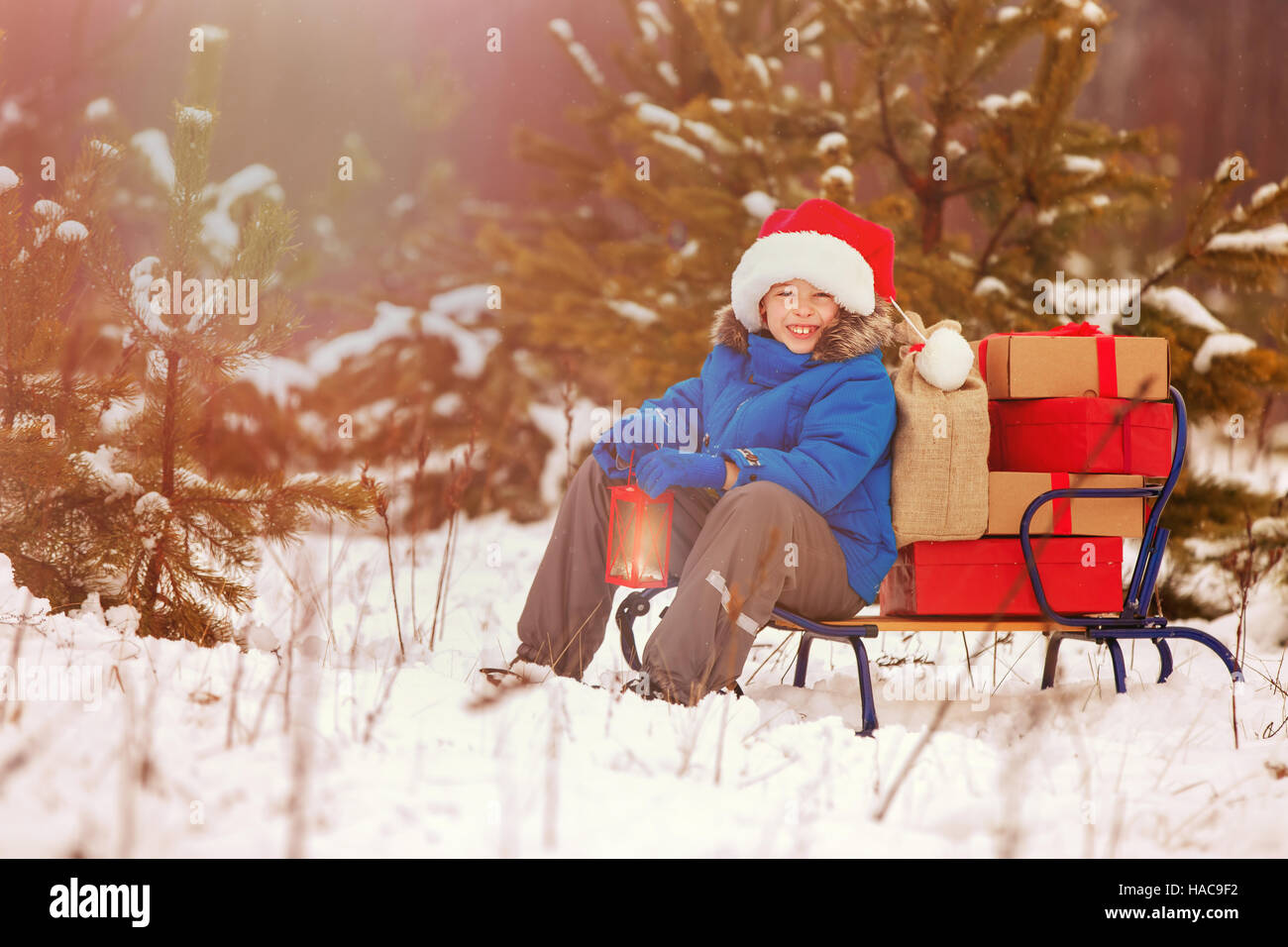 Cute little boy in Santa hat holding Christmas lantern and carries a wooden sled full of gift boxes in snowy forest. Xmas, snow and winter fun for fam Stock Photo