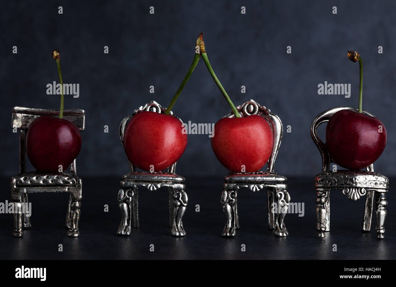 Four red delicious cherries placed on tiny vintage silver chairs on dark background with copy space Stock Photo