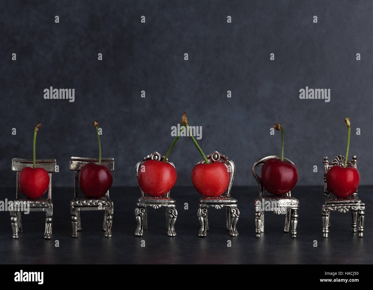 Row of red delicious cherries placed on tiny vintage silver chairs on dark background with copy space Stock Photo