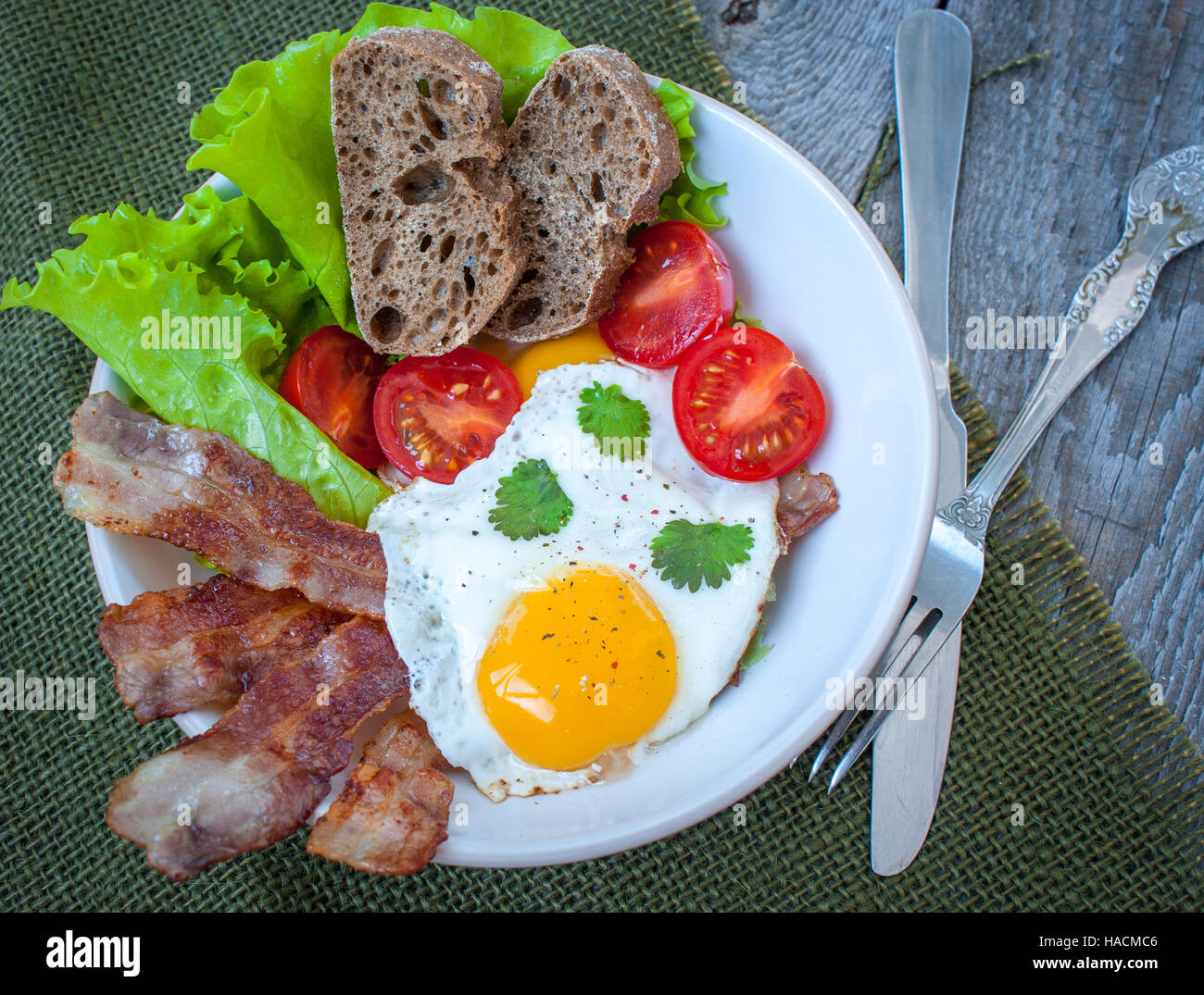 Classic hearty breakfast. Fried egg, bacon, vegetables (lettuce, tomato), greens and bread made from whole wheat flour. Stock Photo