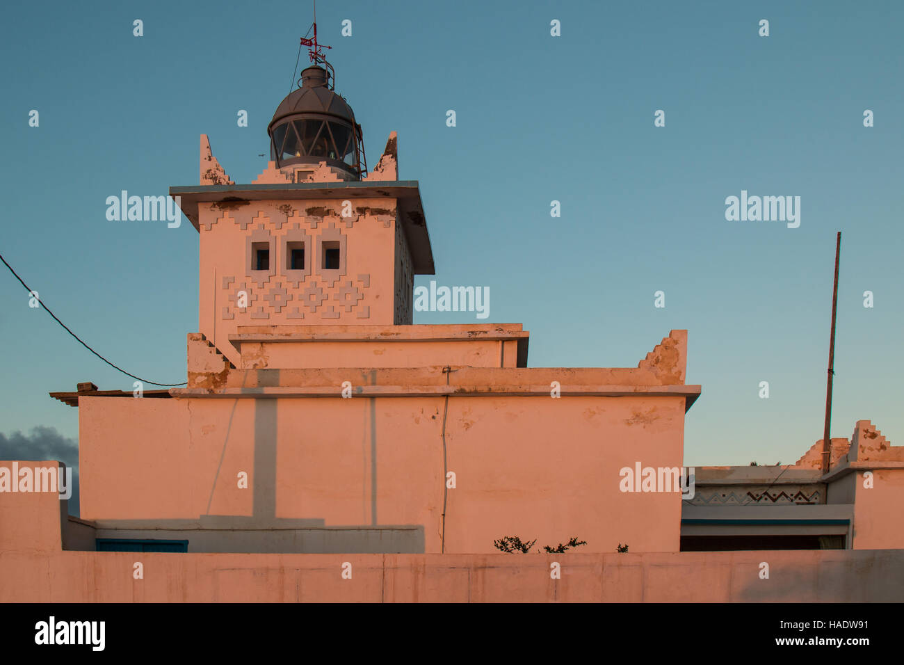 Walls of the lighthouse colored into an orange color by the sunset light. Blue early evening sky. Tower with a light, still off. Sidi Ifni, Morocco. Stock Photo