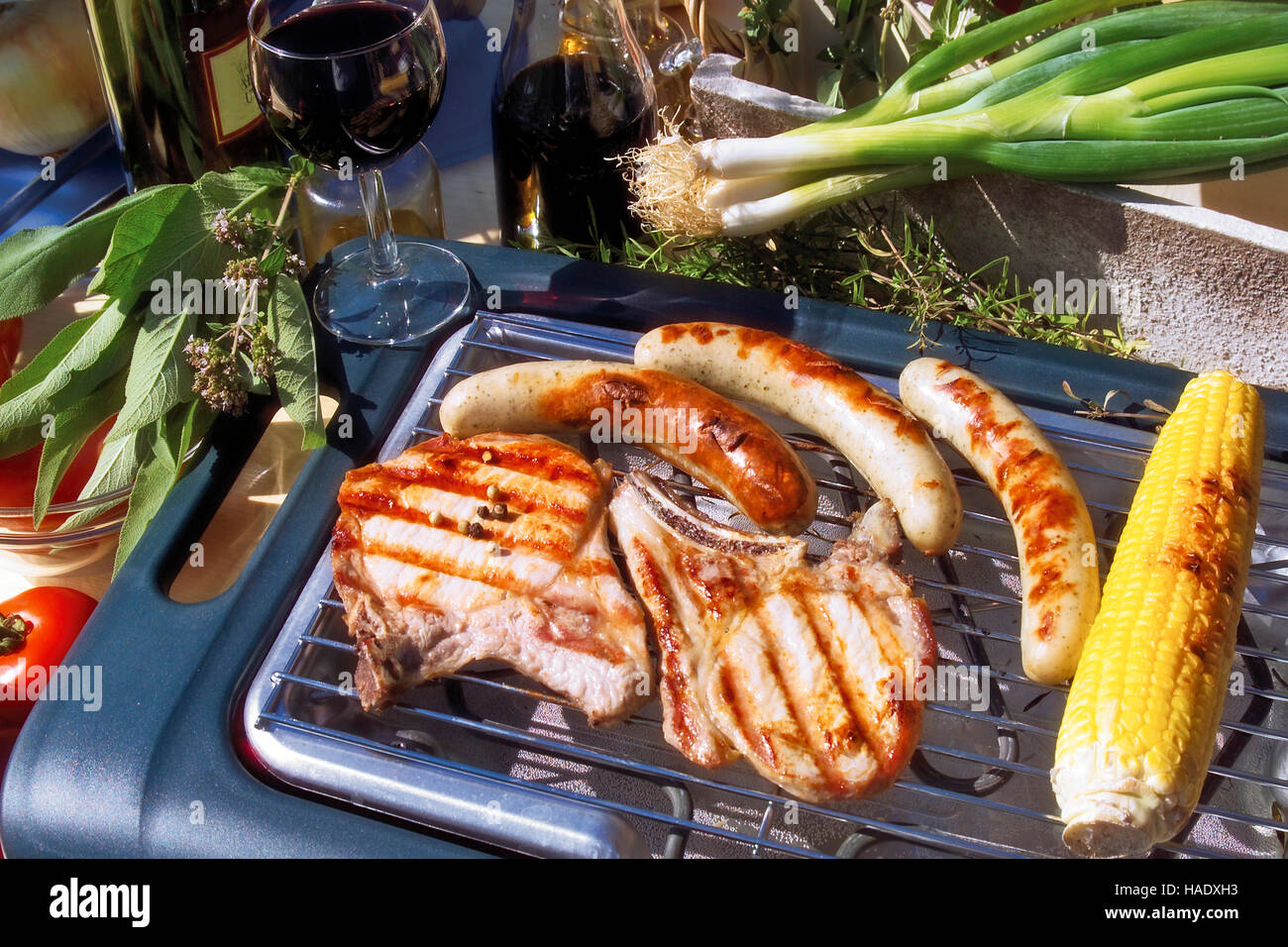 Outdoor barbecue, meat chops, sausages, spring onions, herbs, corn on the cob, red wine Stock Photo