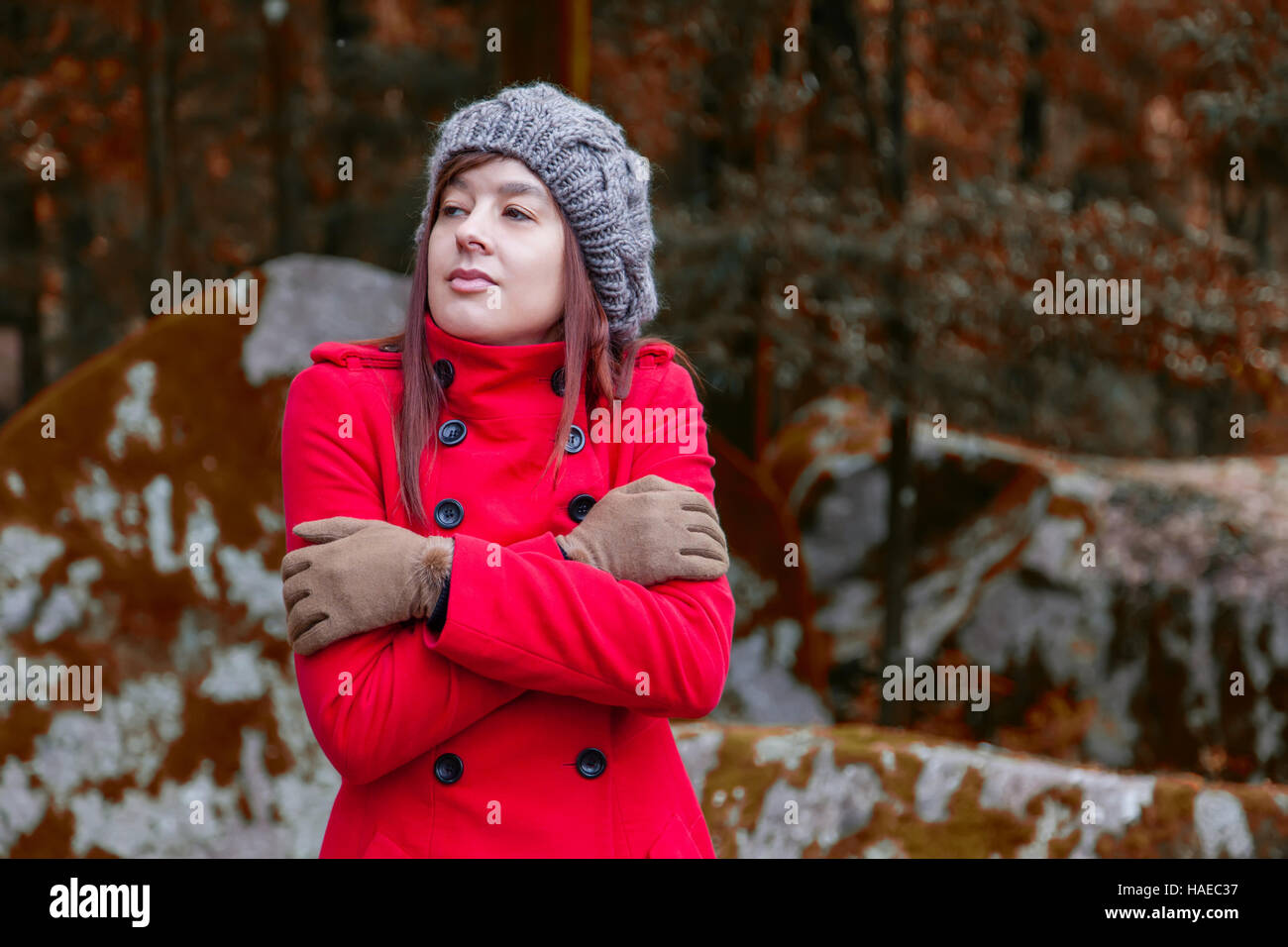 Young woman shivering with cold and embracing herself on a forest wearing a red overcoat, a beanie and gloves during winter Stock Photo