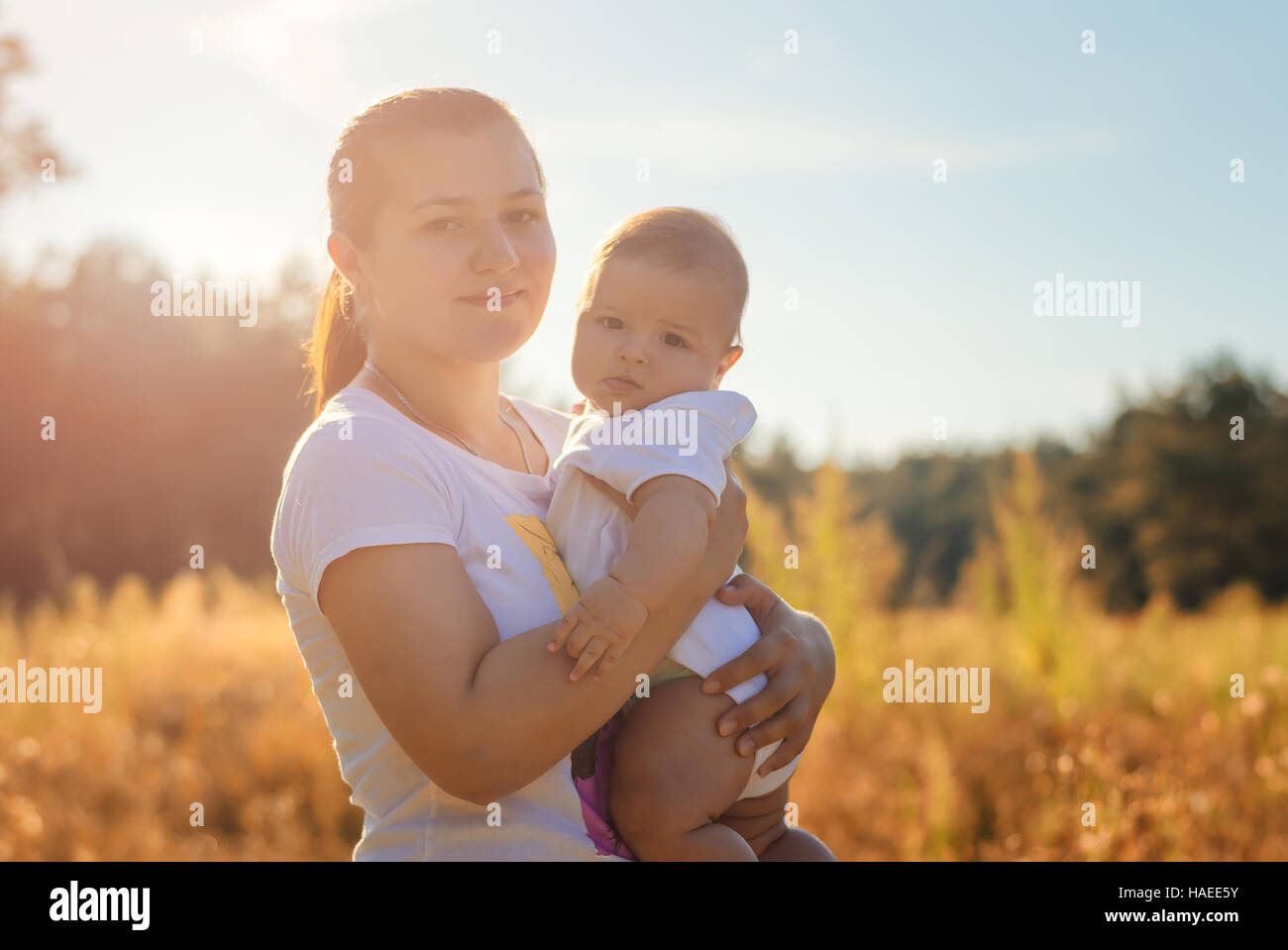 Young beautiful mother holding a baby in her arms. Walk in the autumn warm evening outdoors at sunset. Solar flare lights Woman and child. Stock Photo