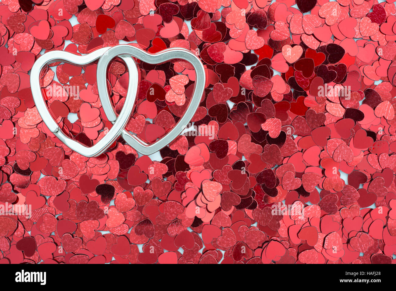 two linked metallic rings in shape of heart on the red confetti background Stock Photo