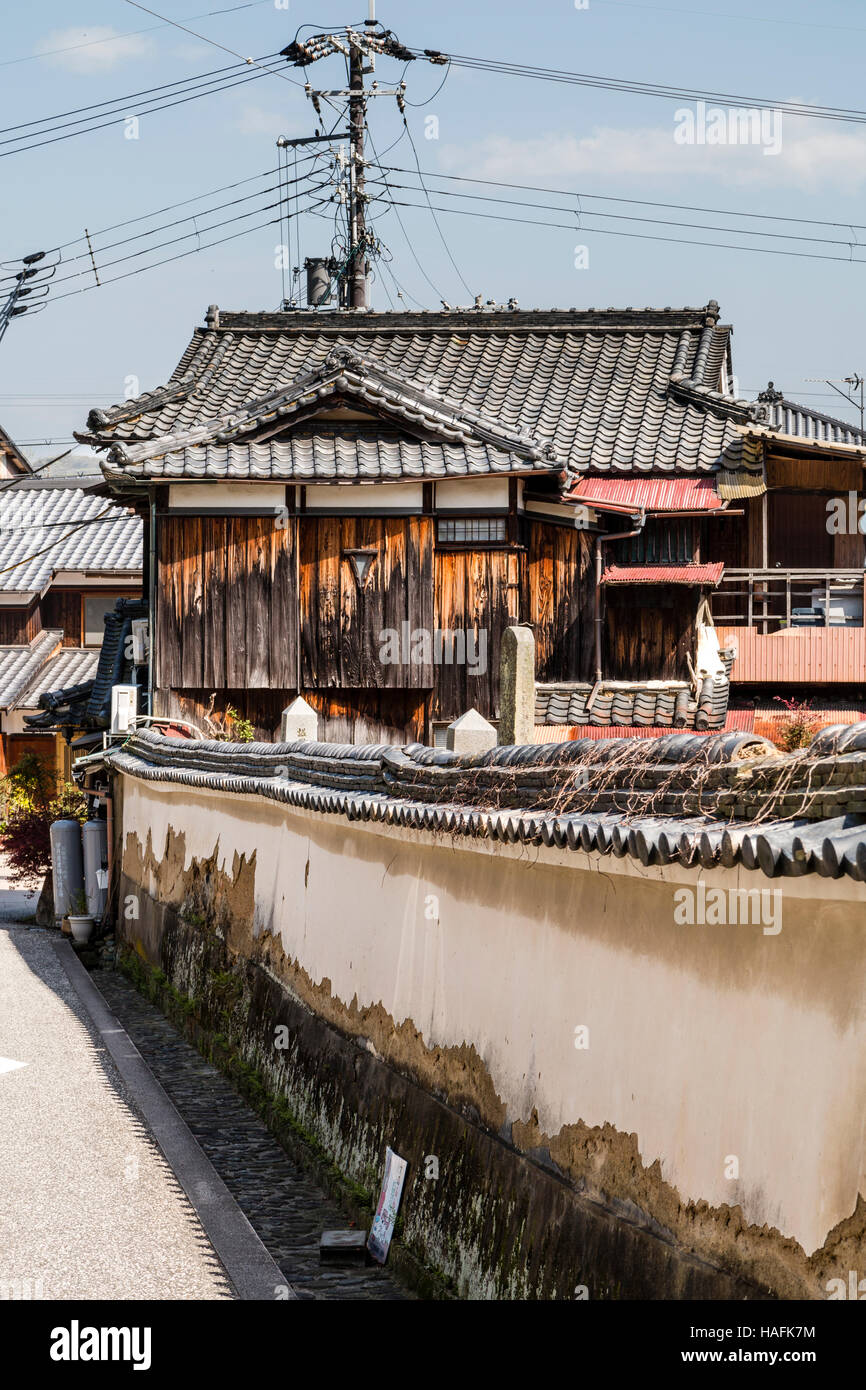 Japan, Tatsuno. Weathered -white plaster roofed wall leading to wooden old style rural Japanese house with small triangular window in the back. Stock Photo