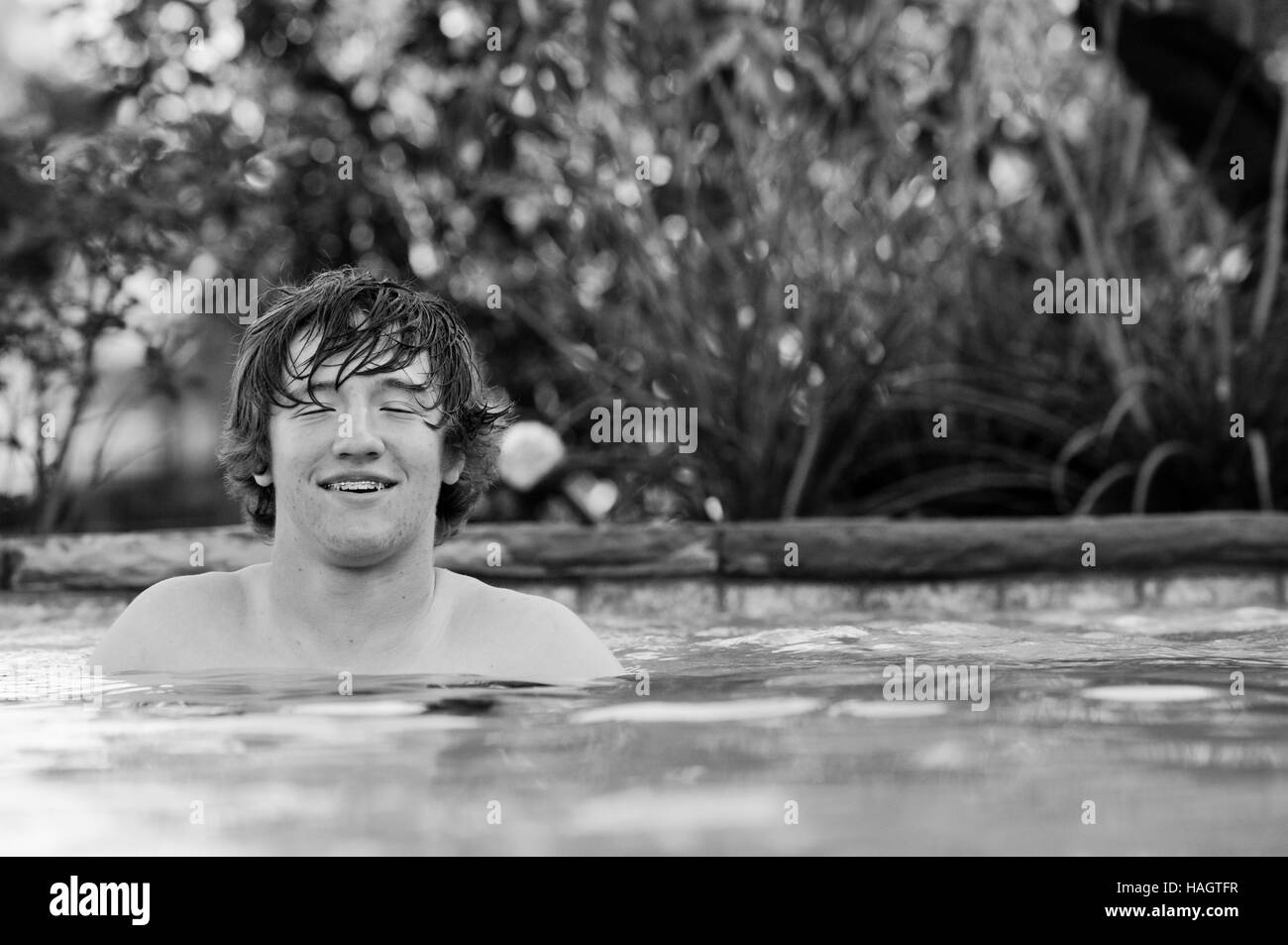 Black and white photo of handsome teenage boy with eyes closed and braces on teeth standing in blue swimming pool. Stock Photo