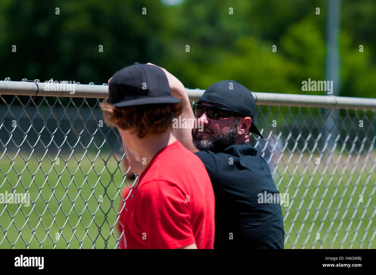 Teen boy and middle-aged father leaning on fence watching baseball game. Stock Photo