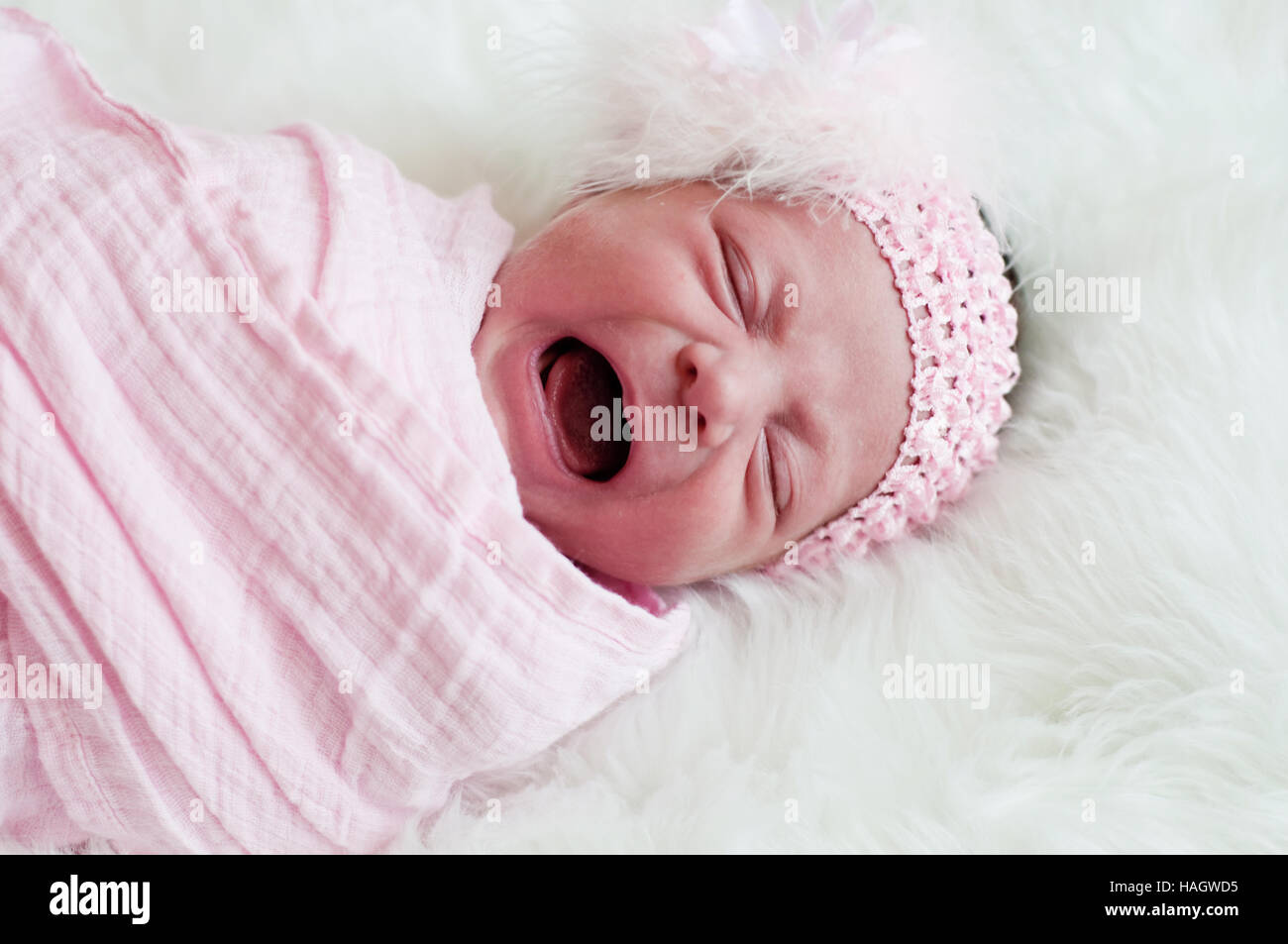 Crying newborn baby girl in pink headband laying on white fur. Stock Photo
