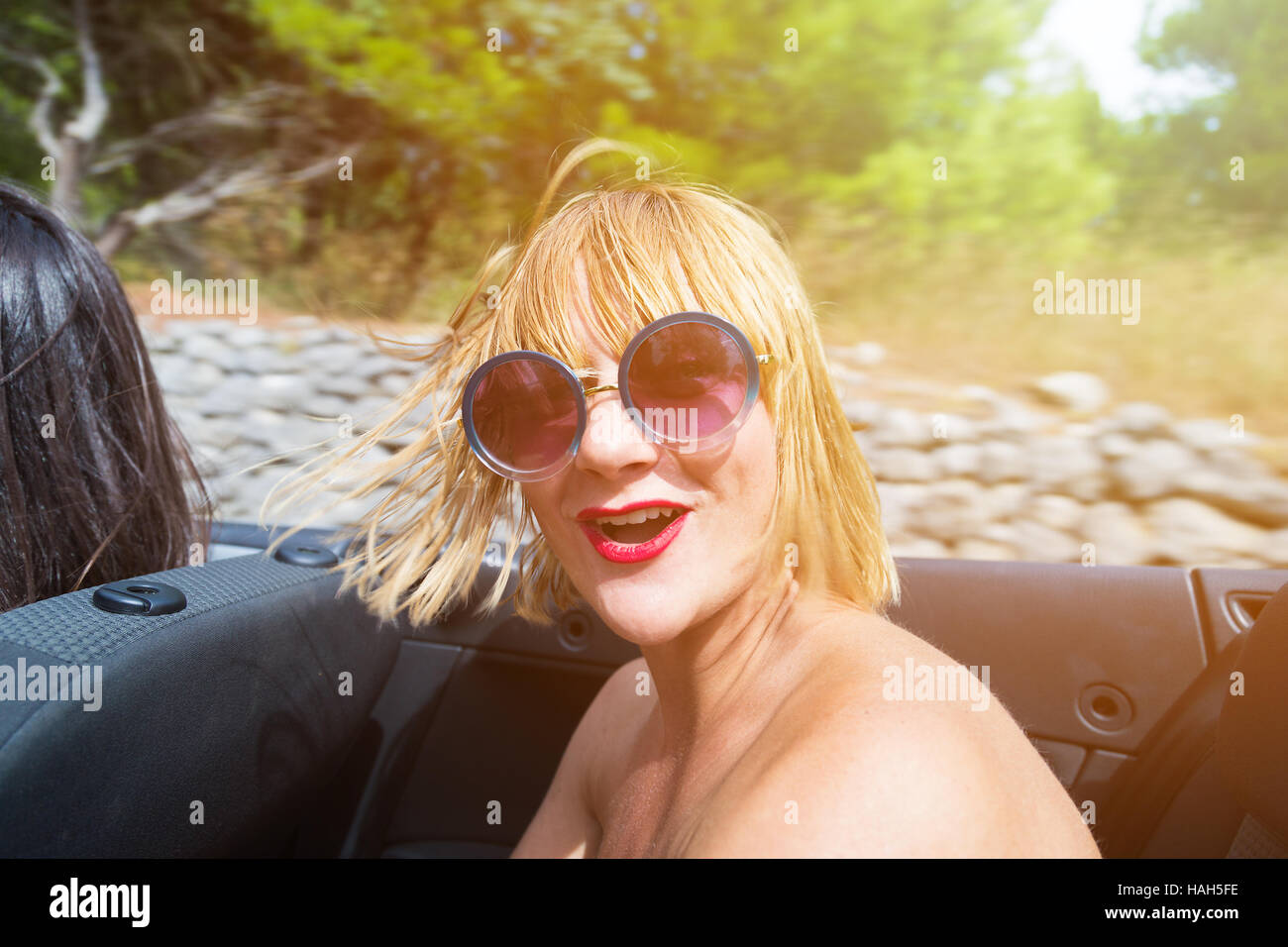 Happy brunette girl sitting and smiling in the back seat of convertible car. Sun effect applied. Stock Photo