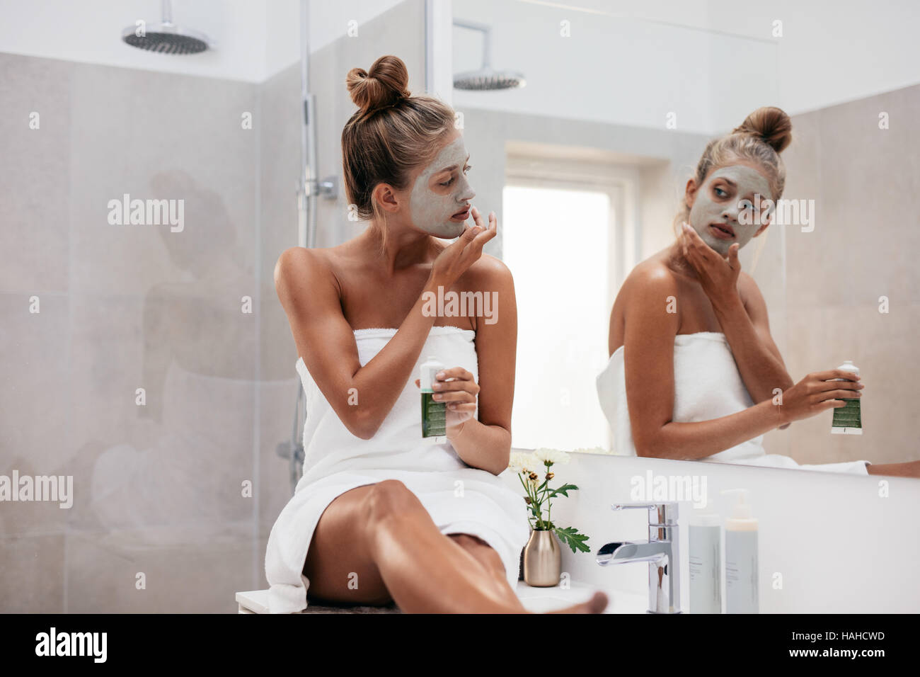 Young woman sitting in bathroom and applying facepack. Beautiful female in front of mirror applying facial mask. Stock Photo
