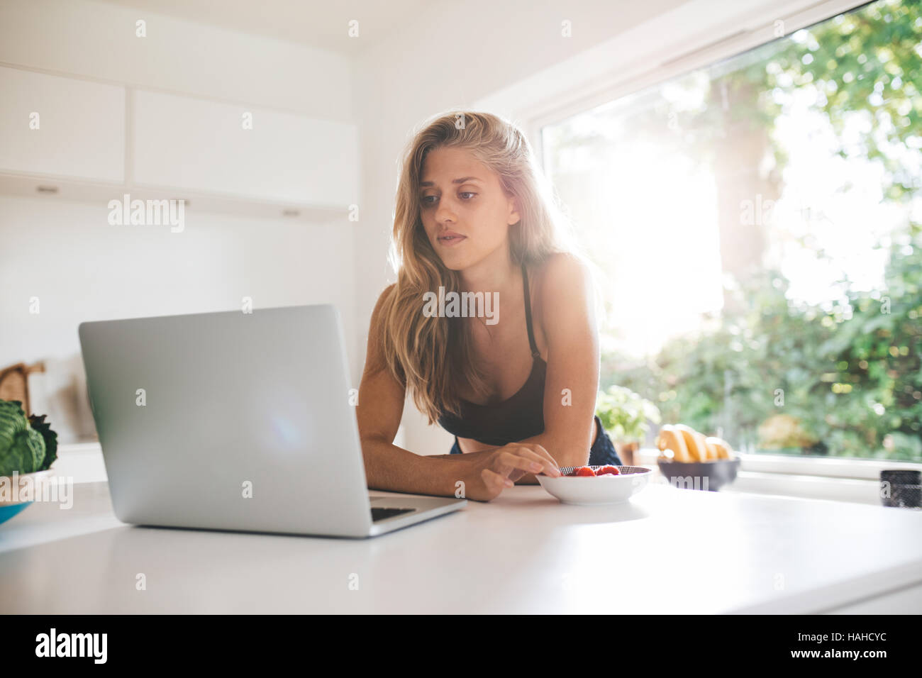 Young woman leaning on kitchen counter and looking at laptop. Beautiful female eating berries and using laptop. Stock Photo