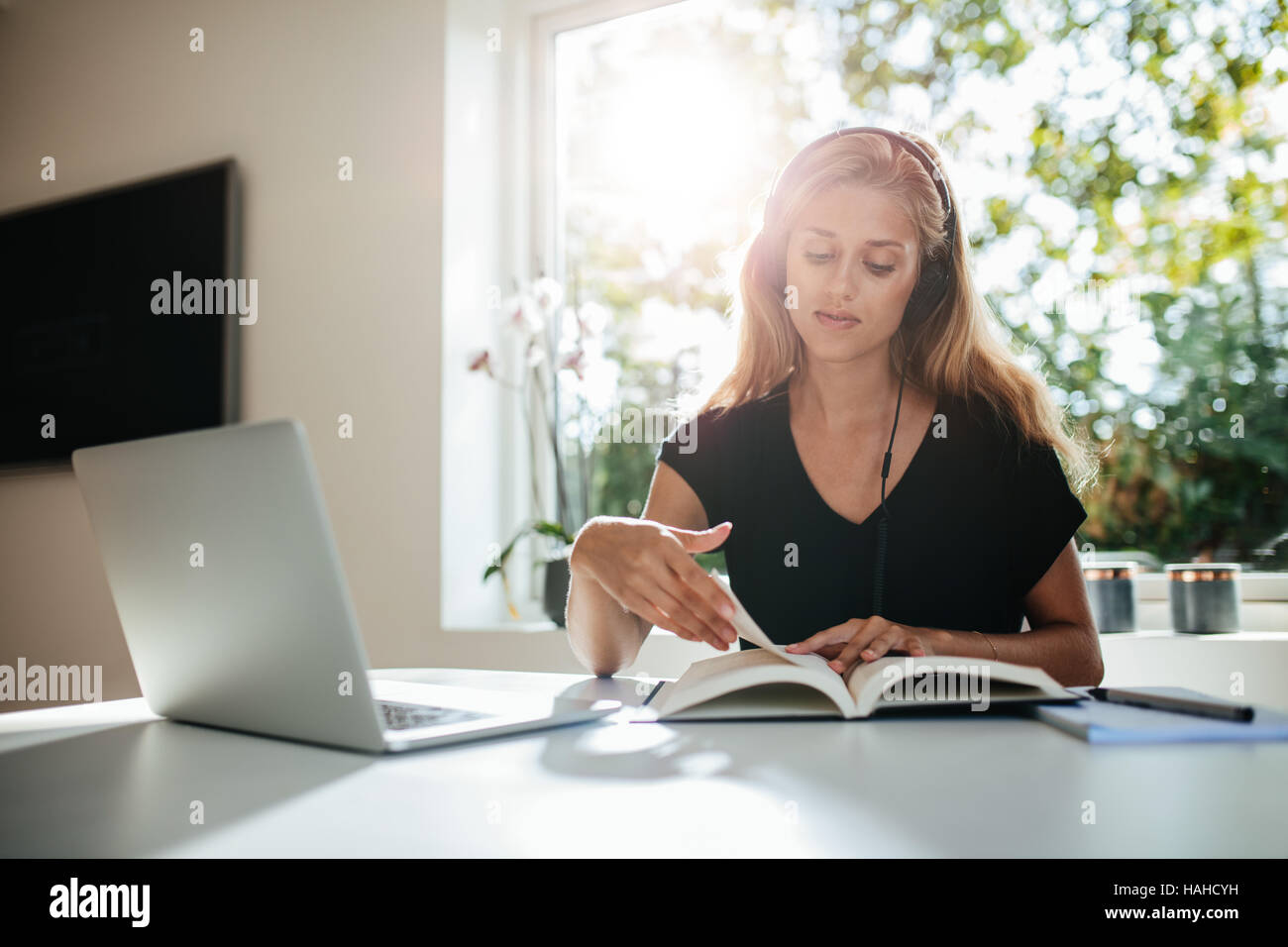Young woman studying at home. Young woman sitting at table with laptop reading book. Stock Photo