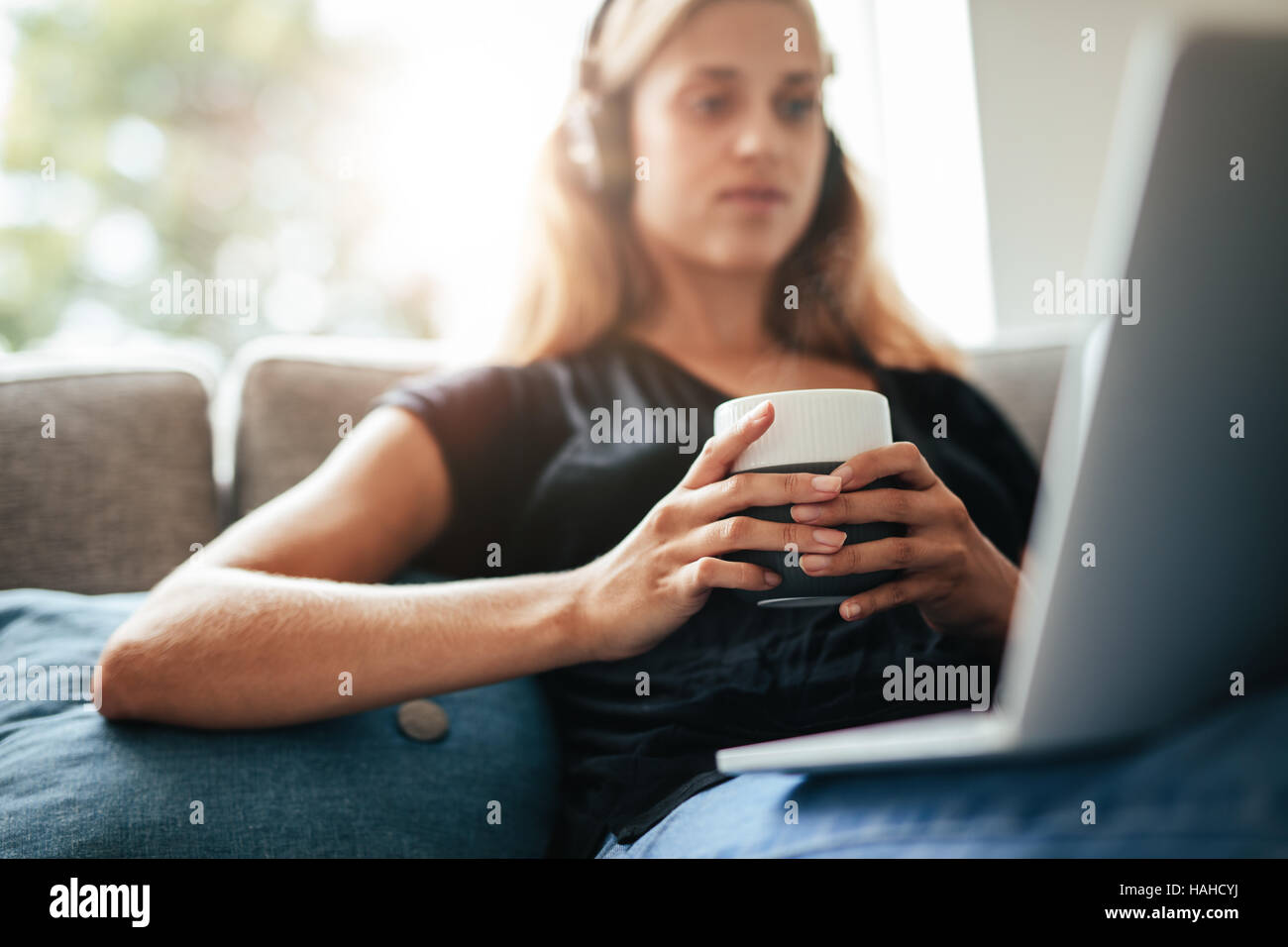 Cup of coffee in hands of a woman relaxing on sofa and looking at laptop. Female relaxing at home. Stock Photo