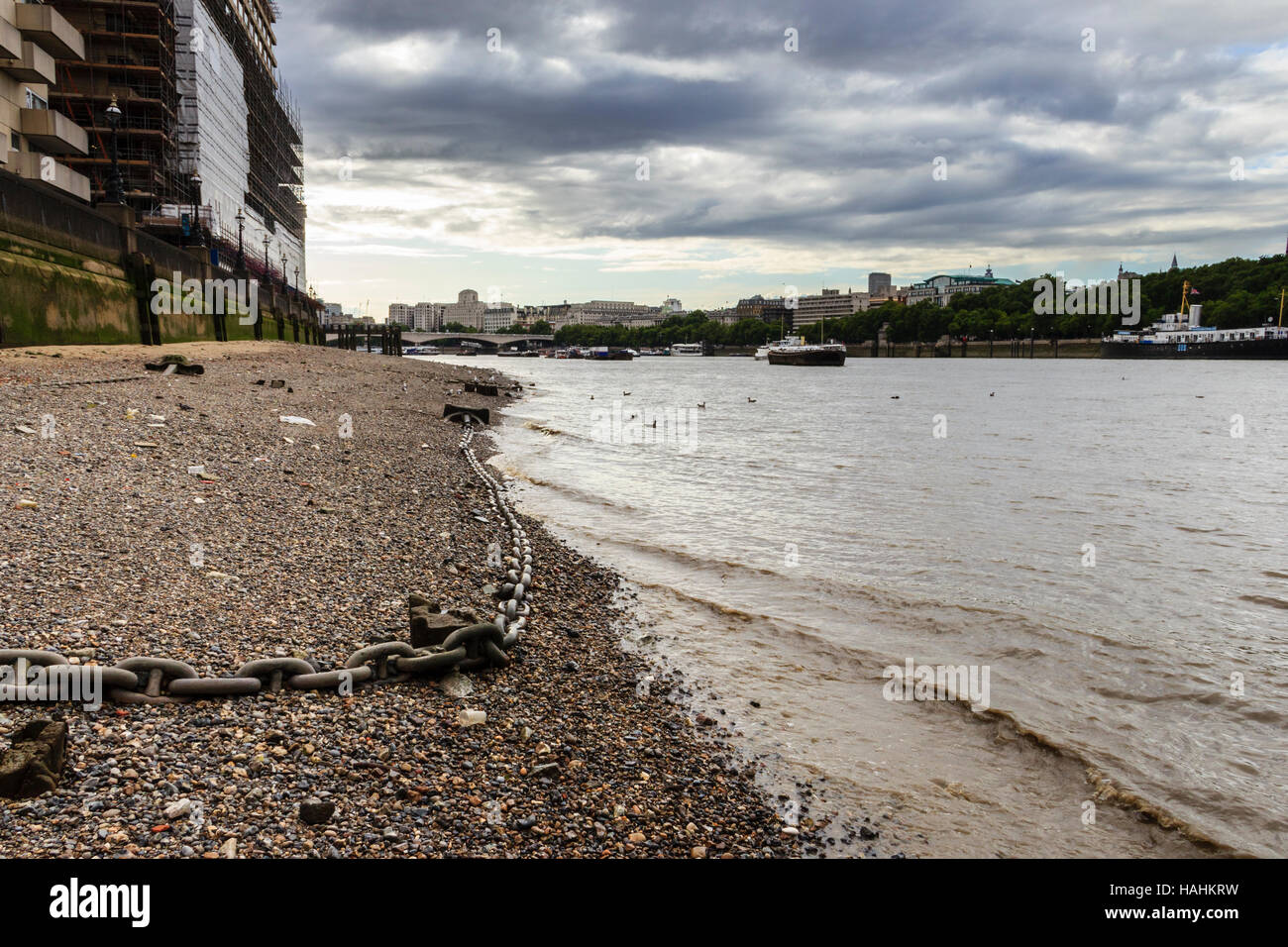 An anchor chain snaking along the stony south shore of the River Thames at Blackfriars, London, UK looking upriver, storm clouds looming overhead Stock Photo