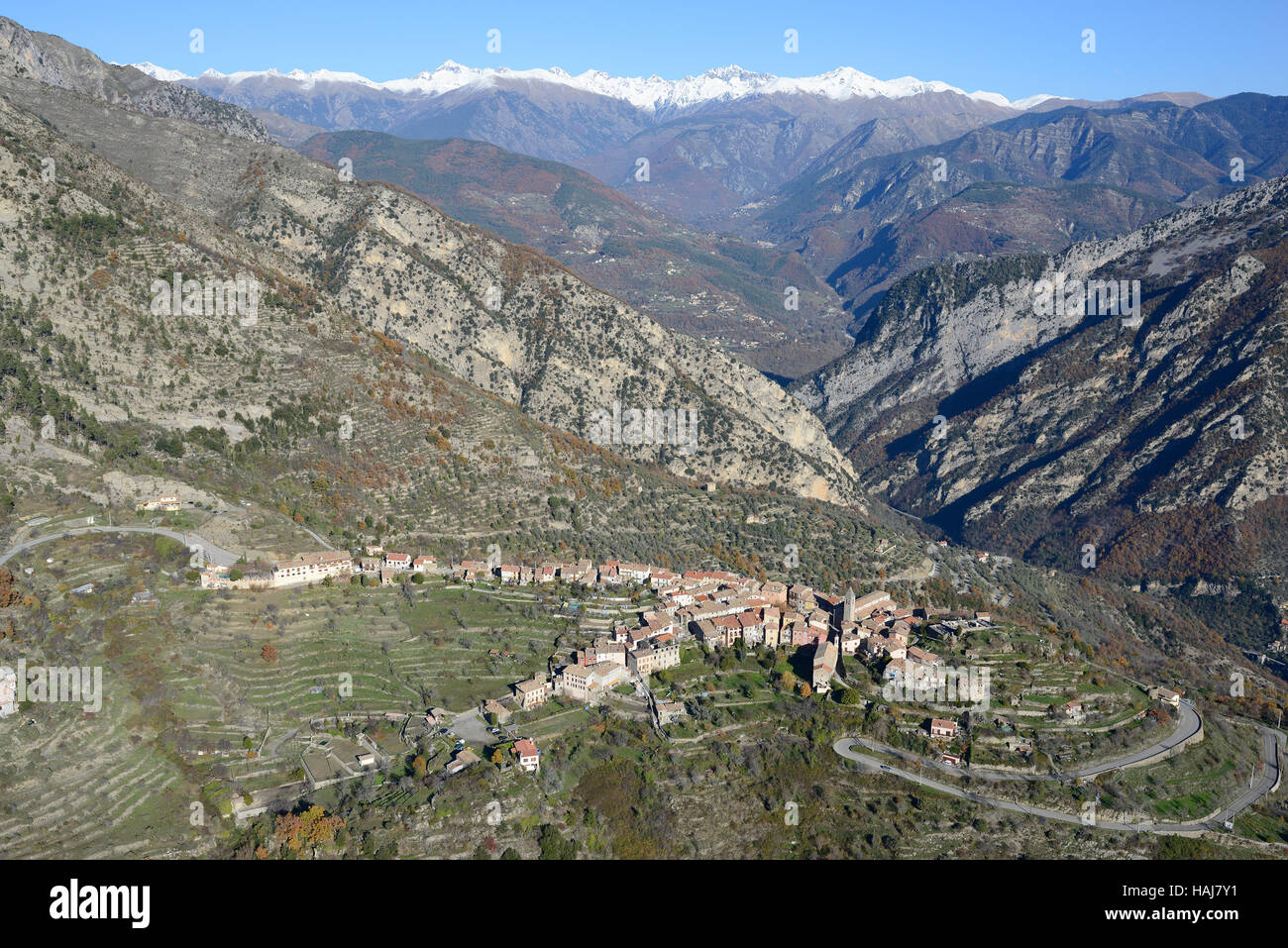 AERIAL VIEW. Hilltop village overlooking the Vésubie Valley with the snowcapped Mercantour Alps in the distance. Utelle, Alpes-Maritimes, France. Stock Photo
